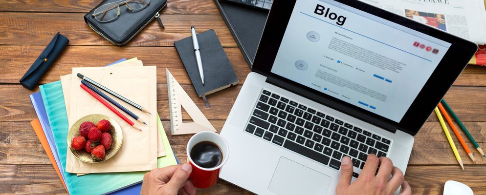 Person looking at a blog on a laptop, drinking coffee, with office supplies scattered.