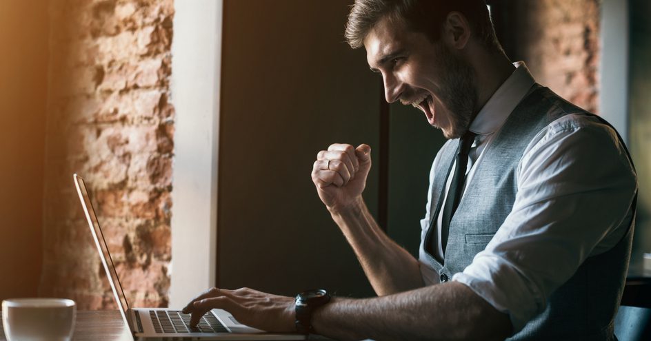 A man at a laptop fist pumping with a cheerful attitude.