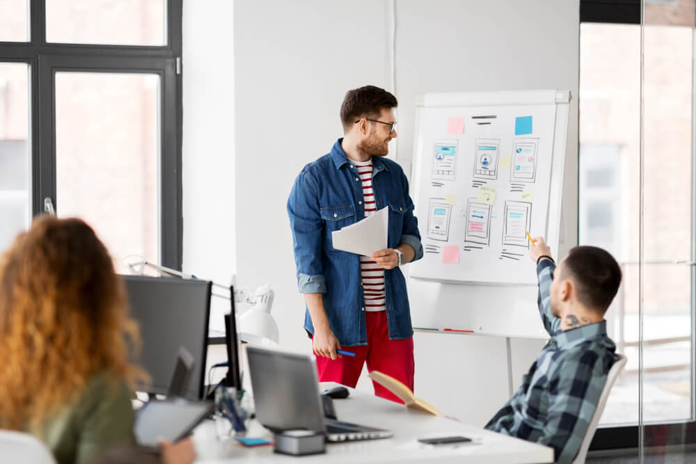 man showing user interface design on flip chart to creative team at office presentation