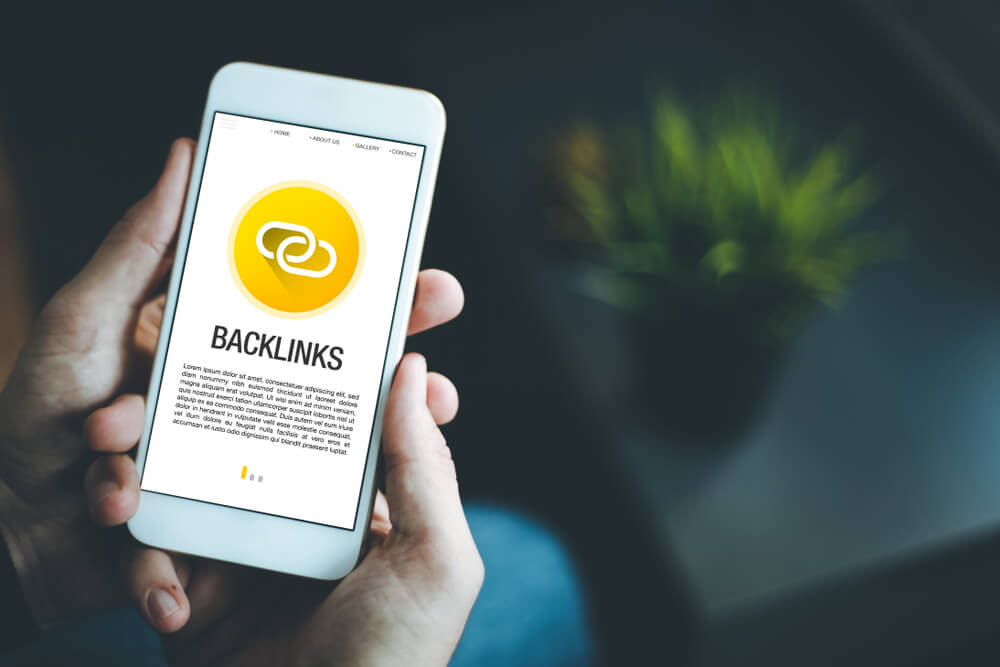 A phone held in two hands shows the word "Local Backlinks" with a yellow backlink icon and small, illegible text. 