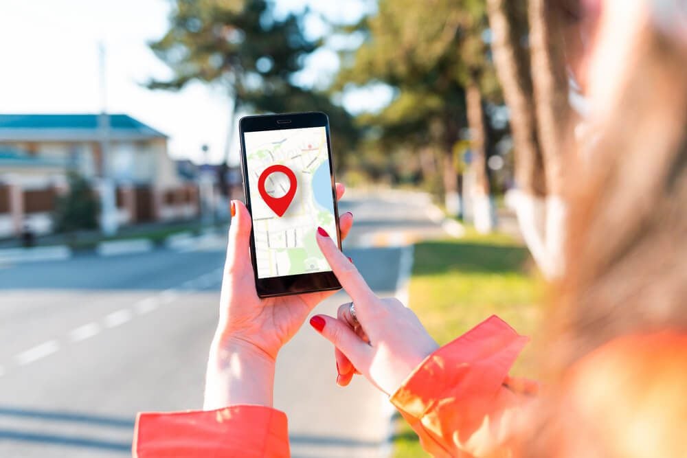 local search_A woman's hand holds a smartphone with an online map that has a red geolocation icon on it. In the background, there is a blurred street.Close up. Concept of online navigation and GPS