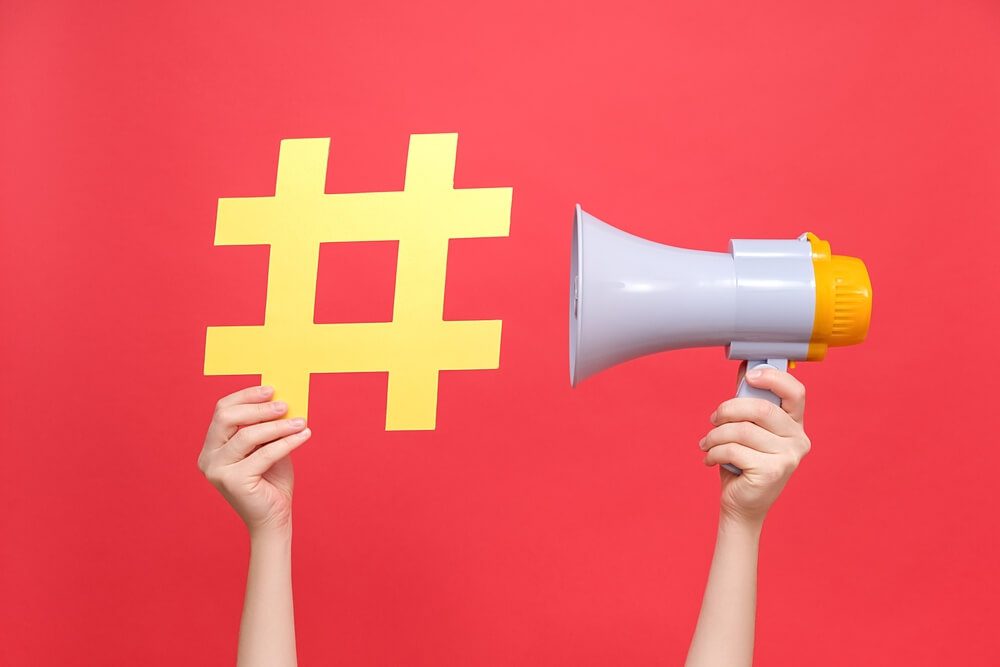 seo topic_Close up female hands holding yellow hashtag sign and megaphone, label for business, marketing and advertising, isolated on red studio background. Concept social network monitoring, media measurement