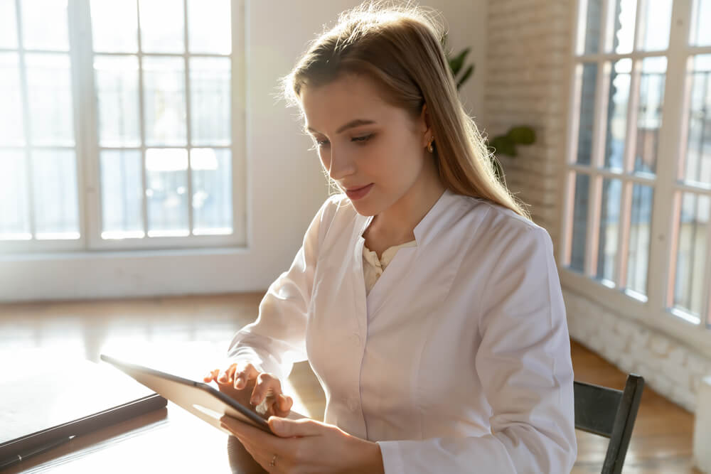 medical search_Focused young woman doctor wearing uniform working with medical checkup results on tablet in office, using gadget, browsing medical apps, chatting, consulting patient online, telemedicine concept