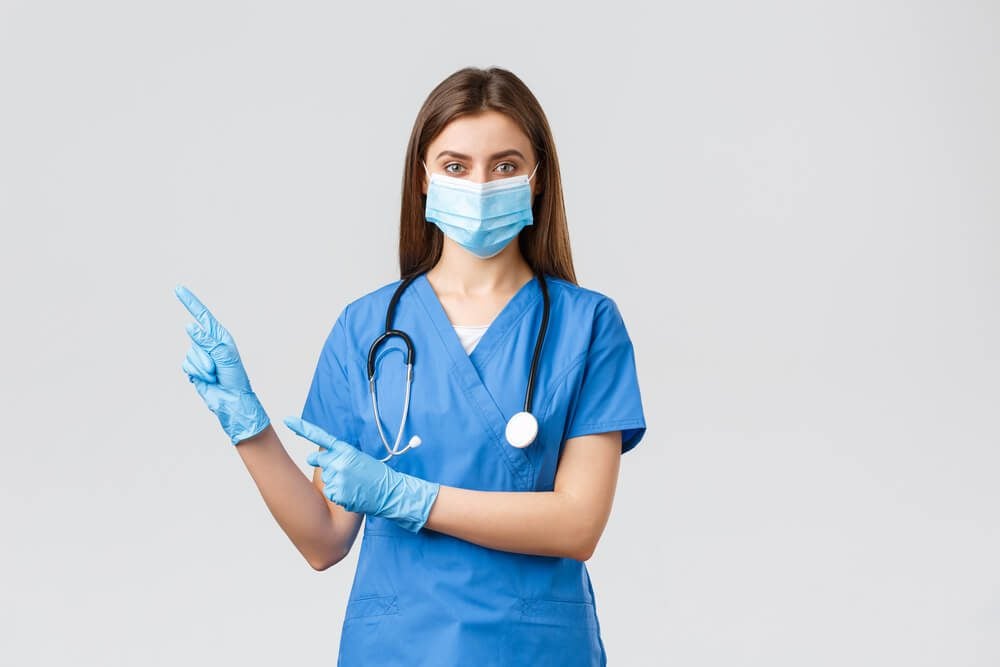 healthcare click_Covid-19, preventing virus, health, healthcare workers and quarantine concept. Pleasant female doctor or nurse in blue scrubs, medical mask and gloves, invite follow page or click link, pointing left