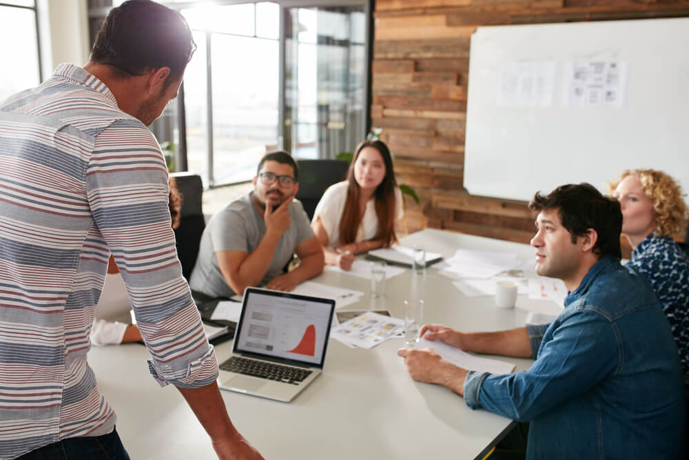 agency_Young man giving business presentation on laptop to colleagues sitting around table in conference room.