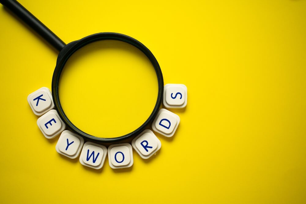 A magnifying glass and the word "keywords" made with letter game blocks surrounding it, on a bright yellow background. Conceptual image of the importance of the keywords in searching engines.