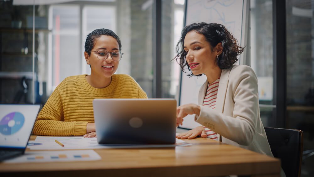 agency_Two Diverse Multiethnic Female Have a Discussion in Meeting Room Behind Glass Walls in an Agency. Creative Director and Project Manager Compare Business Results on Laptop and App Designs in an Office.