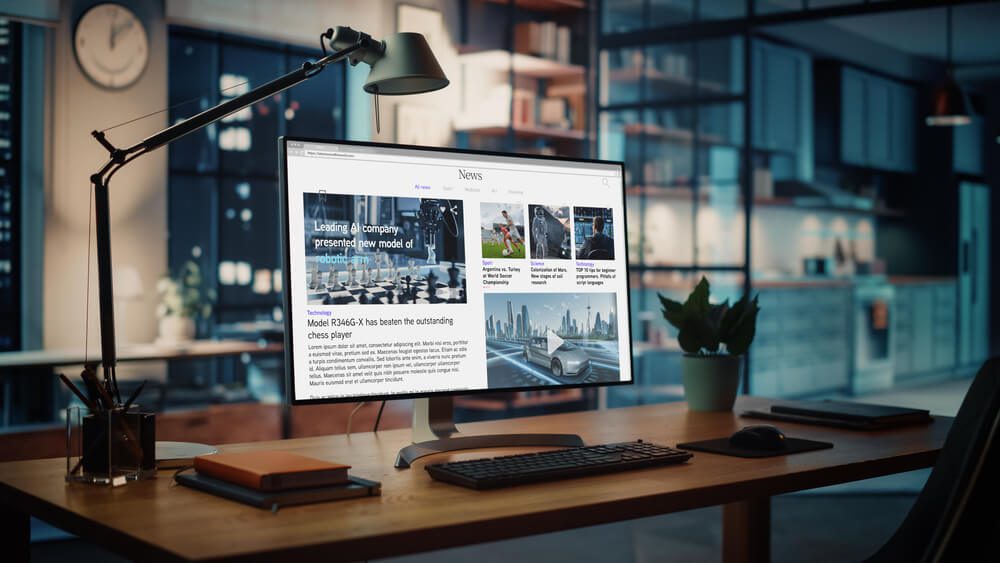 website page_Shot of a Desktop Computer with Latest News Web Page Showing On Screen Standing on the Wooden Desk in the Creative Cozy Living Room. In the Background Warm Evening Lighting and Open Space Studio.