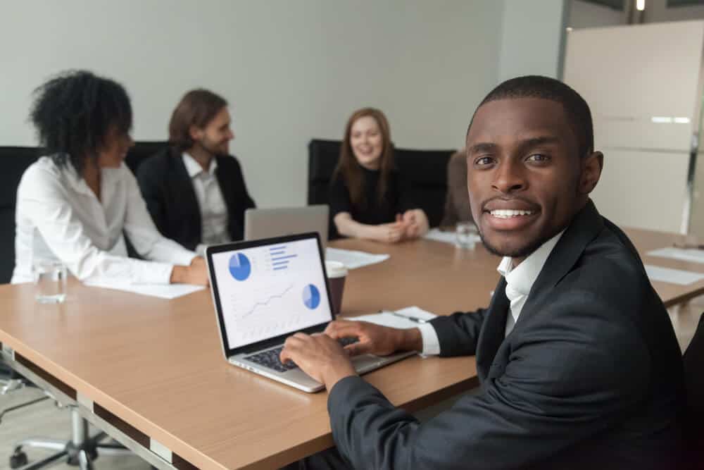 seo partner_Smiling african-american businessman in suit looking at camera at diverse team meeting, business analyst using laptop with project statistics at screen, professional black manager at work portrait