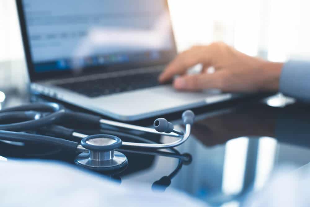 medical websites_Male doctor hand working on laptop computer, browsing internet or medical research with stethoscope on desk in doctor office, close up. Medical and technology concept, close up