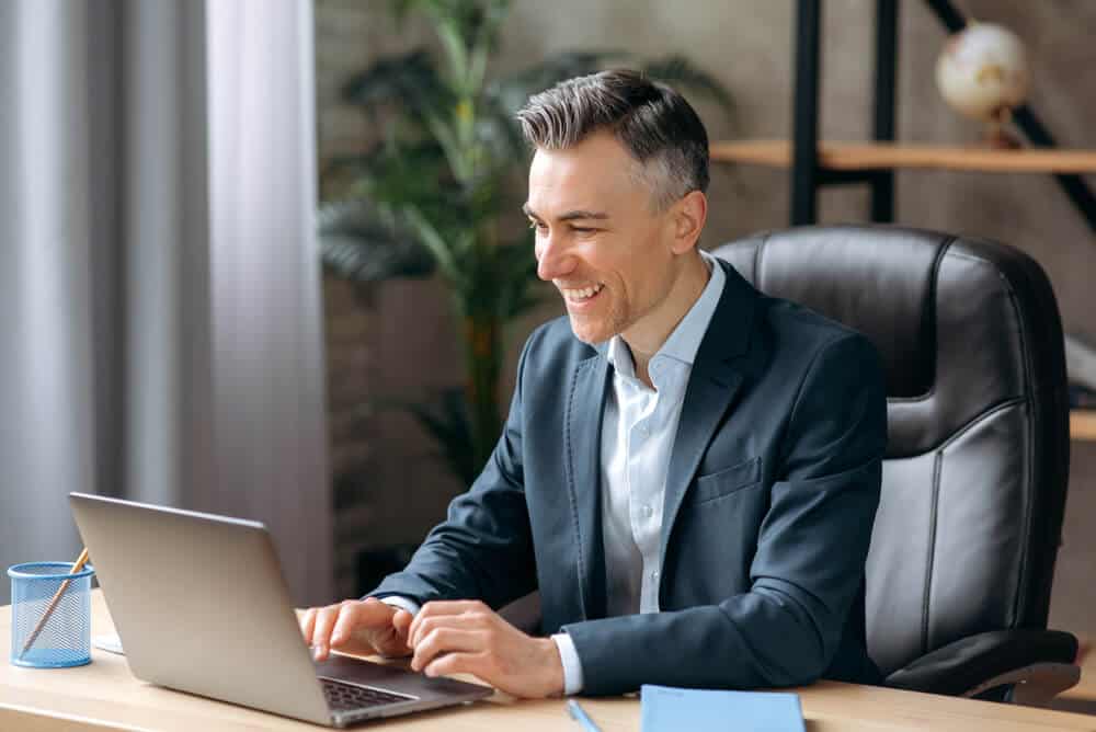 seo friendly_Influential successful Caucasian adult businessman or manager in a formal suit uses a laptop, communicates via video conferencing, online meetings, sits office, types on a keyboard, smiles friendly