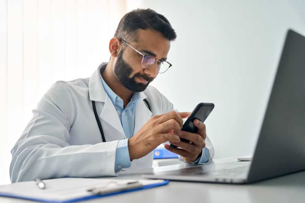 therapist website_Indian male serious doctor medical worker in modern clinic wearing eyeglasses and white coat uniform using cell mobile smartphone apps, laptop computer. Medicine technologies health care concept.