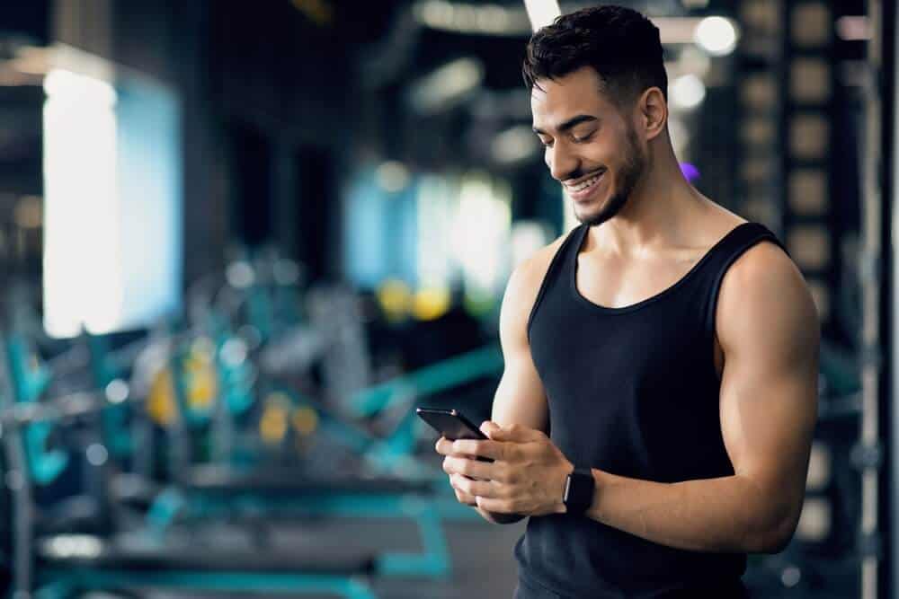 gyms website_Handsome Young Middle Eastern Man Using Smartphone During Training In Sport Club, Smiling Arab Male Athlete Messaging On Cellphone Or Browsing New Fitness App While Spending Time At Gym, Copy Space