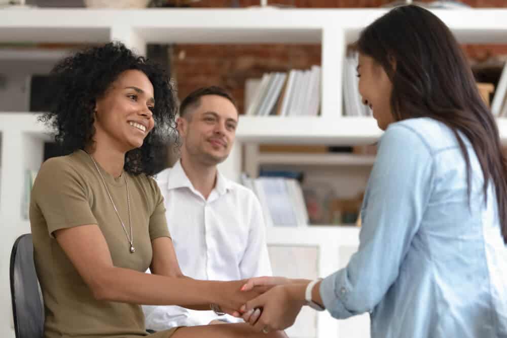 drug rehab center_Asian and african women holding hands during group therapy session, diverse friends feeling reconciled relief smiling giving psychological support empathy overcome problem at psychotherapy counseling