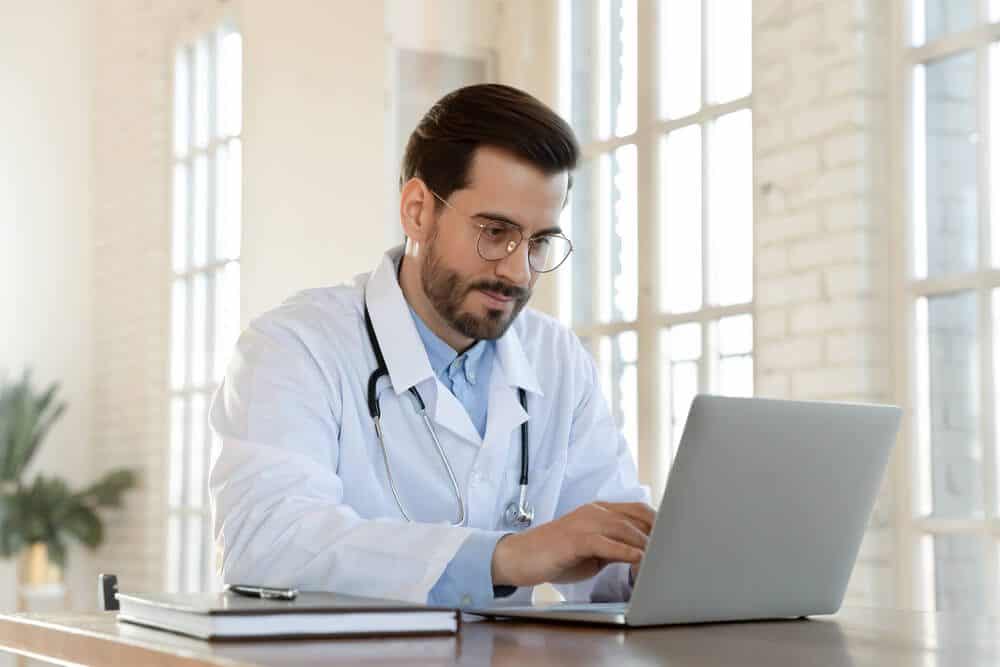 healthcare email_Focused young doctor wearing glasses working on laptop in office, sitting at desk, looking at screen, serious therapist gp writing report or email, consulting patient online, filling medical card