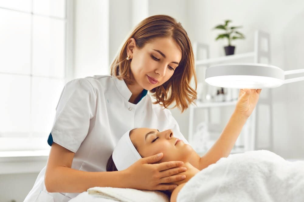 dermatologists_Young woman cosmetologist or dermatologist directing lamp for facial treatment to lying young woman in beauty spa salon. Facial treatment, massage, skincare, cosmetology concept