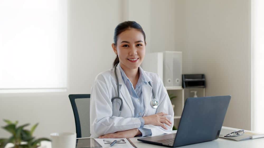 dermatologist_Young adult asia people or medic staff woman sitting toothy smile arm cross look at camera happy work on desk in clinic office room in telehealth telemedicine smart health care consult online service.