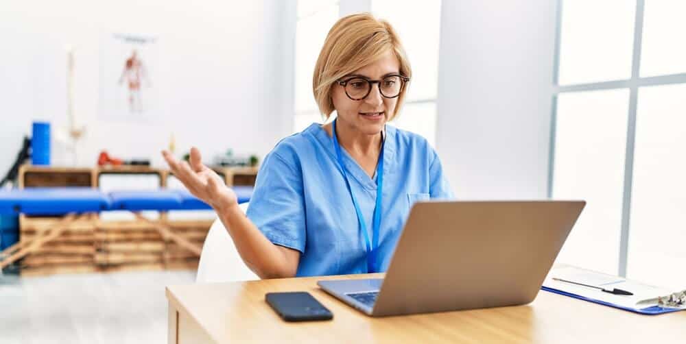 rehab center online_Middle age blonde woman wearing physio therapy uniform having video call at clinic