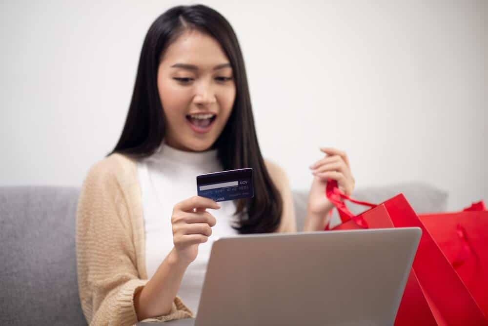 A woman sits on a couch with her laptop on her lap. She's holding a credit card in one hand and two red shopping bags in the other hand.