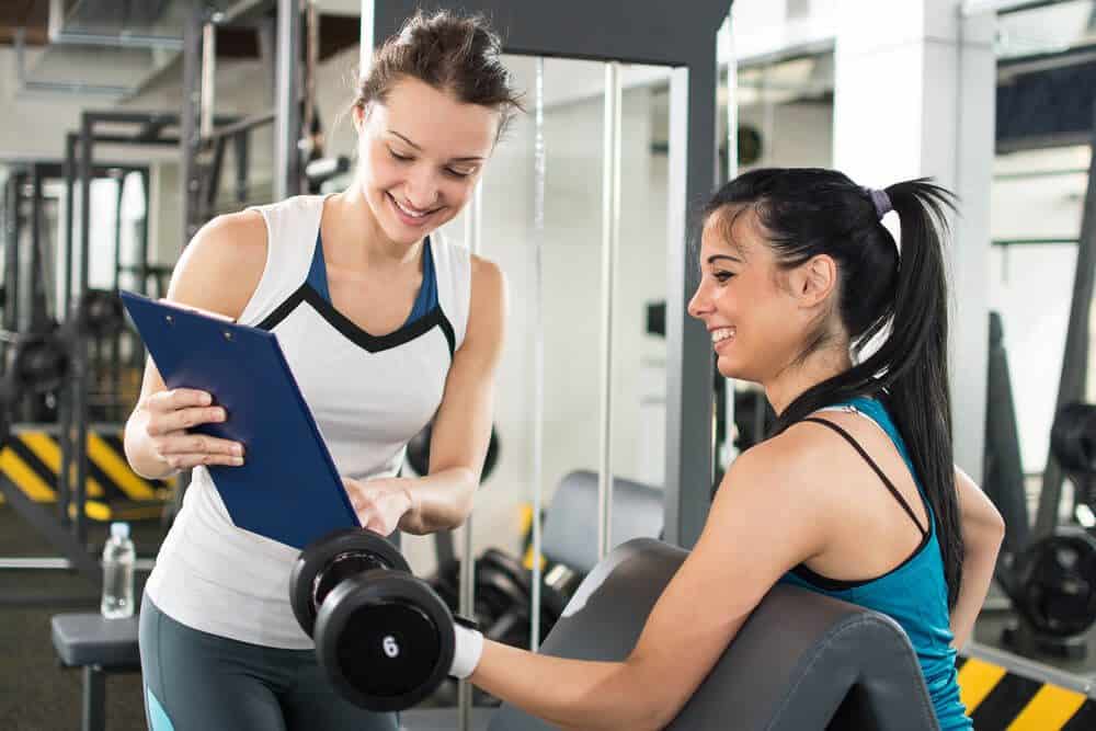 personal trainer_Female personal trainer helping young woman on her work out routines in gym. Fitness woman lifting weights and her personal trainer showing fitness report on a clipboard.