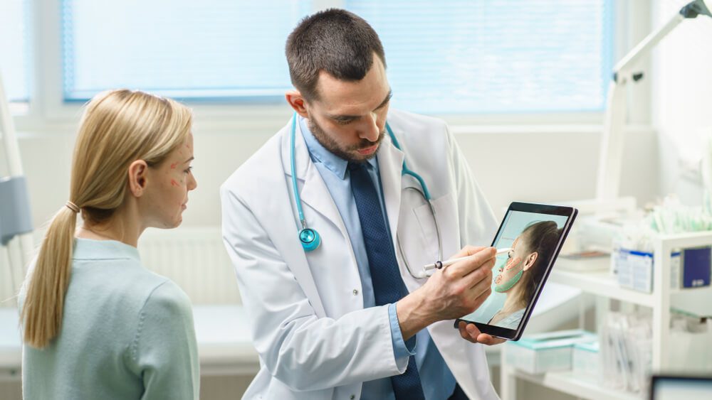 plastic surgeon_Plastic / Cosmetic Surgeon Consults Woman about Facial Lift Surgery, He Draws Arrows on Digital Tablet Computer Screen, Showing Types of Facelift and Nose Correcting Procedures Available for Her.