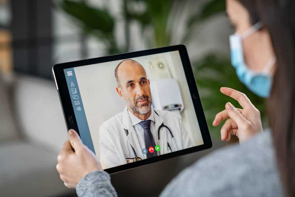 online medical_Sick woman wearing protective mask during video call with mature doctor. Back view of patient with surgical face mask talking during conference call with her physician, staying at home in quarantine.