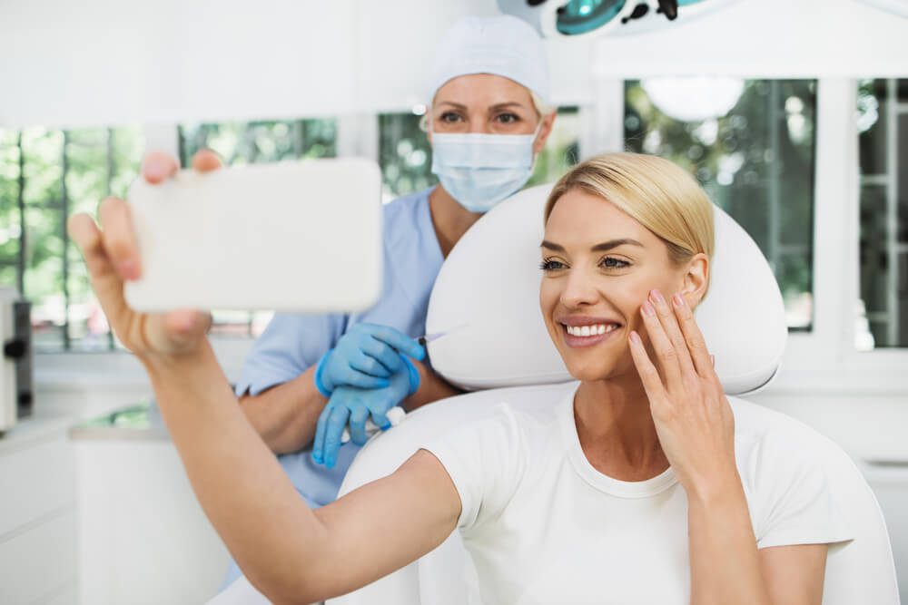 plastic surgeon_Beautiful and happy blonde woman at beauty medical clinic. She is sitting and talking with female doctor about face esthetics treatment.