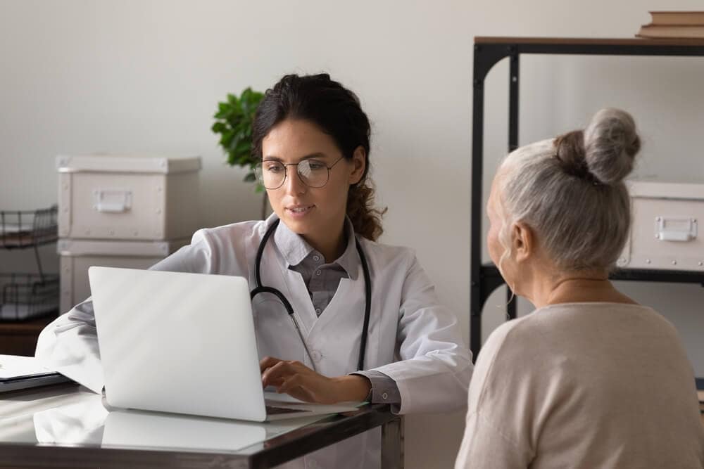 healthcare patient_Serious young female doctor meeting with elderly patient in office, listening to woman health problems complaints, typing on laptop, keeping electronic database on computer. Geriatric medic care