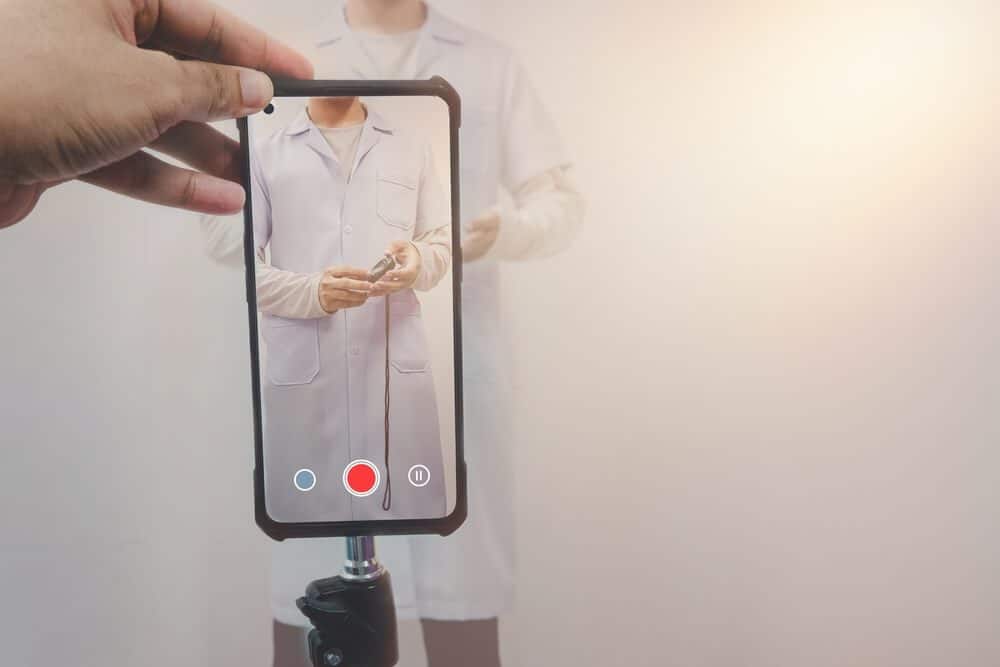 healthcare content_Selective focus on smartphone sharing on social media by asian male doctor live broadcasting education for healthcare viral video clip on white-gray background. Telemedicine or telehealth concept.