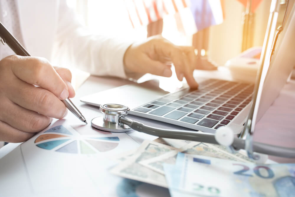 hospital investment_Doctor working on laptop computer with report analysis and money about Healthcare costs and fees in medical hostpital office. Focus stethoscope on table. Healthcare budget and business concept