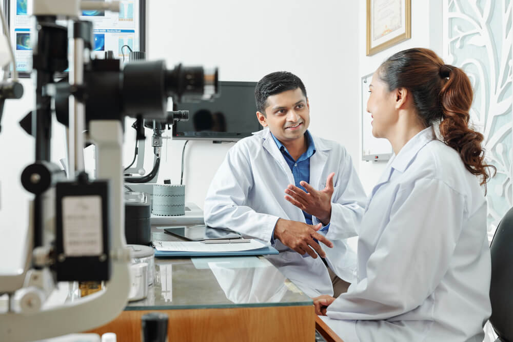 optometry_Indian male optometrist in white coat sitting and discussing with his nurse some working moments at office