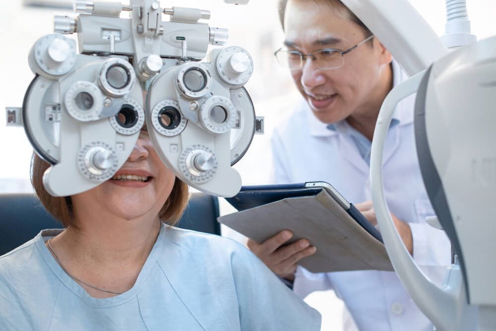 optometrist_Senior Asian woman looking through optical phoropter during eye exam,with an optometrist nearby, diagnostic ophthalmology equipment, selective focus