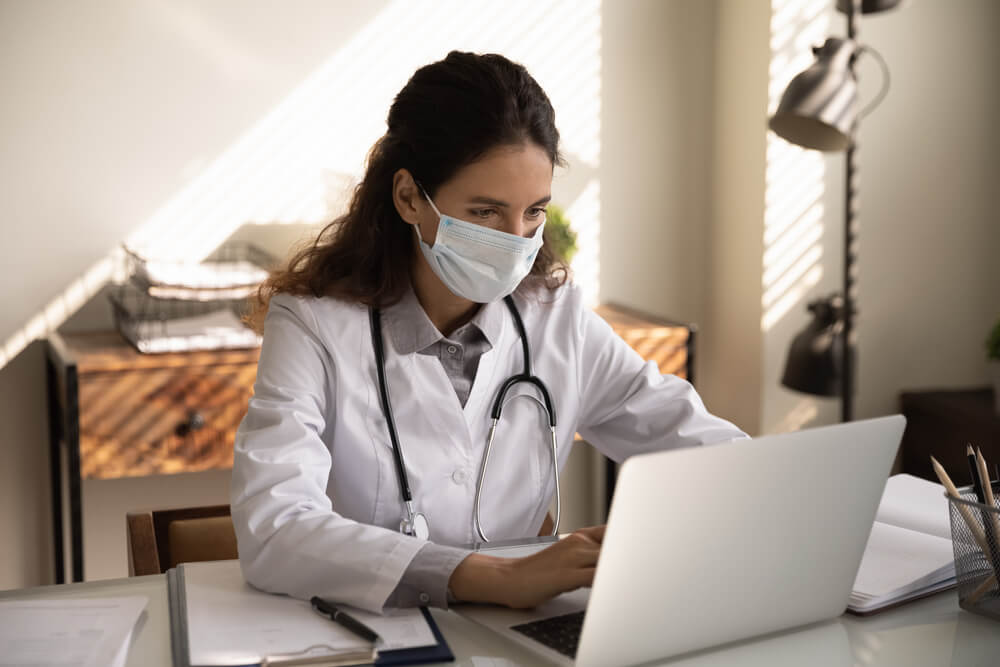 healthcare email_Close up focused professional female doctor physician wearing protective face mask using laptop, looking at screen, consulting patient online, chatting, browsing medical apps, searching information