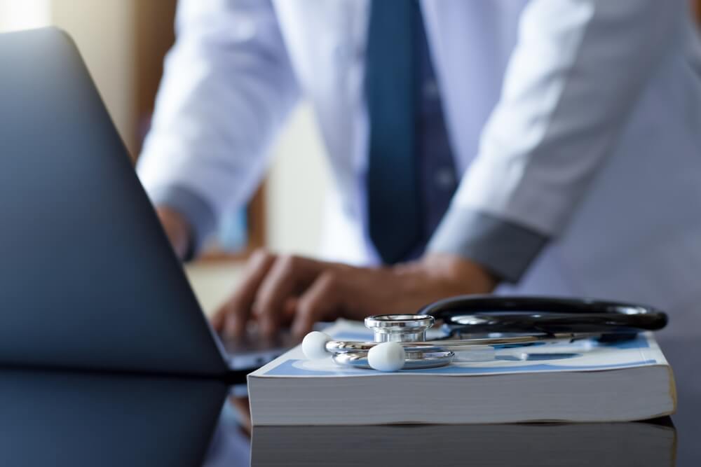 medical email_Male doctor hands typing on laptop computer keyboard with textbook and medical stethoscope on the desk at office. Online medical,medic tech, emr, ehr concept.