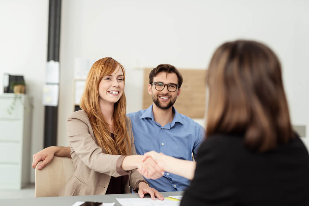 customers_Happy young couple meeting with a broker in her office leaning over the desk to shake hands, view from behind the female agent