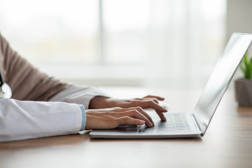 Improving Healthcare Email Marketing Open Rates (9 Proven Tips) 2 healthcare email_Cropped of woman doctor having online training, sitting in her cabinet, using laptop, typing on keyboard, side view, copy space. Unrecognizable lady physician working with computer, clinic interior