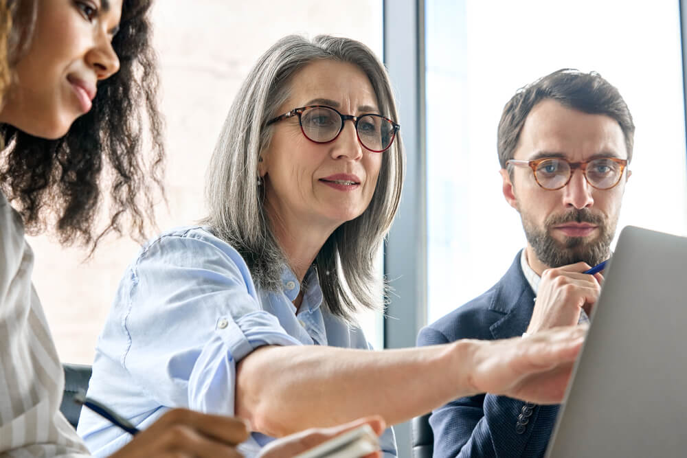 digital partner_Mature older ceo businesswoman mentor in glasses negotiating growth business plan with diverse executive managers at boardroom meeting table using laptop. Multicultural team work together in