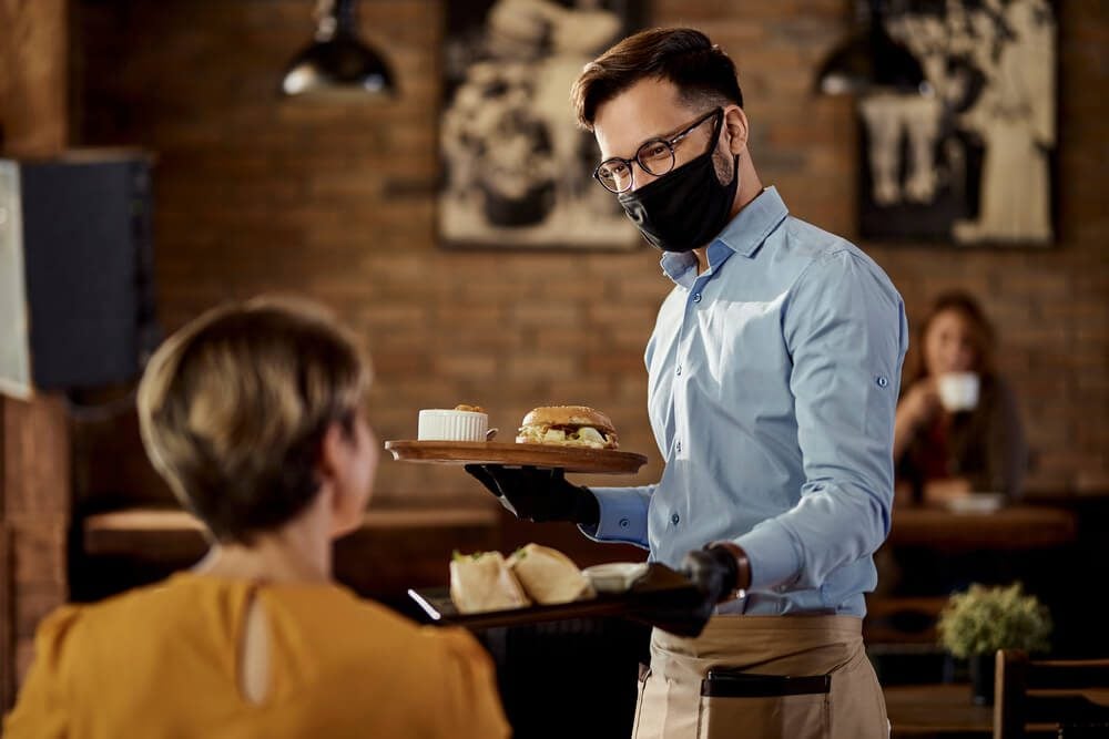 ethnic restaurant_Happy waiter wearing protective face mask and gloves while bringing food to a customer in a pub.