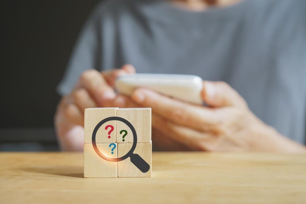 seo questions_magnifying glass with question mark inside on wooden cube block with blurred senior woman holding smart phone