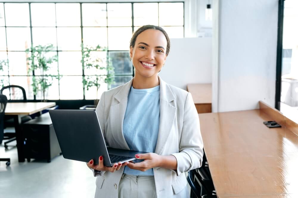 seo partner_Gorgeous confident successful hispanic or brazilian woman, in an elegant clothes, seo, top manager, programmer, stands in a modern office, holding an open laptop in hands, looks at camera, smiling