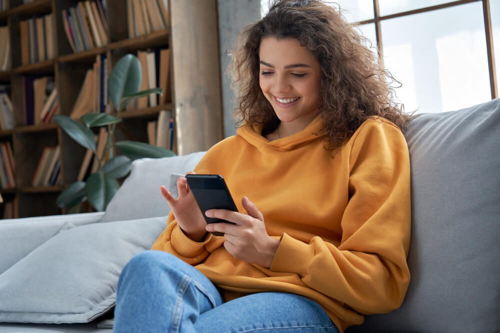 mobile app_Happy millennial hispanic teen girl checking social media holding smartphone at home. Smiling young latin woman using mobile phone app playing game, shopping online, ordering delivery relax on sofa.