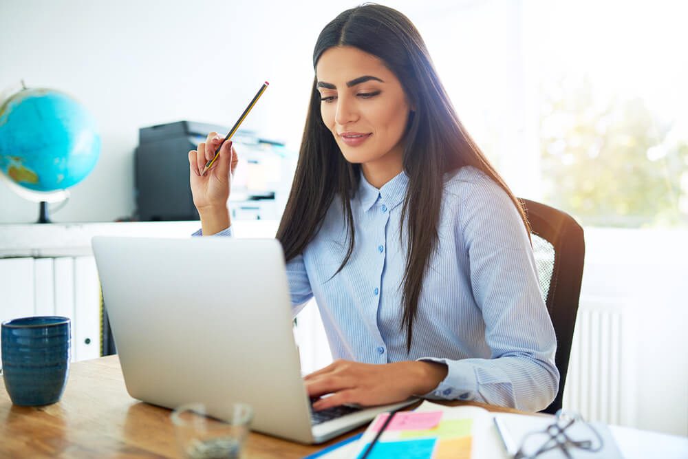 small business_Single calm young Indian woman in blue blouse and long hair holding pencil in hand while seated at desk in front of laptop computer in bright room