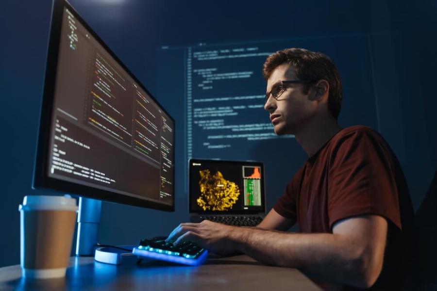 AI Web Development_Side view portrait of modern IT developer using computer at home office at night, working on software, coding new application, sitting in front of big pc monitor. Data science specialist at work