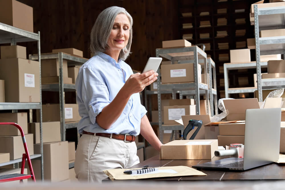 mobile app small business_Female mature senior small business owner using mobile app checking parcel box. Older entrepreneur seller holding cell phone tech device preparing retail package postal shipping order in warehouse.