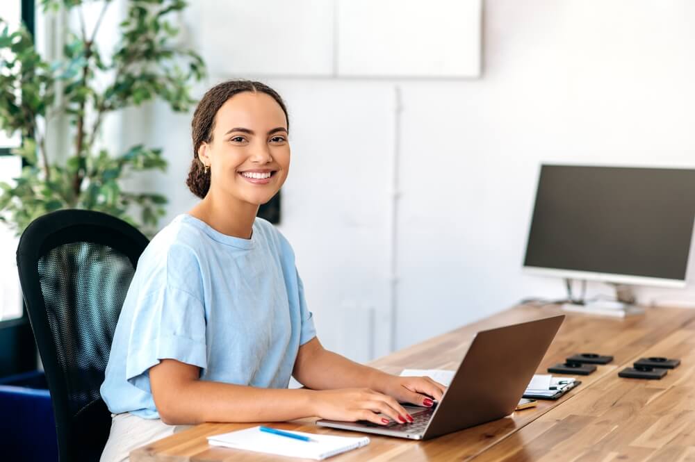 local seo partner_Photo of a successful confident gorgeous mixed race woman, business mentor, financial advisor, programmer, seo, sitting at a work desk in a modern office, looking at camera, smiling friendly