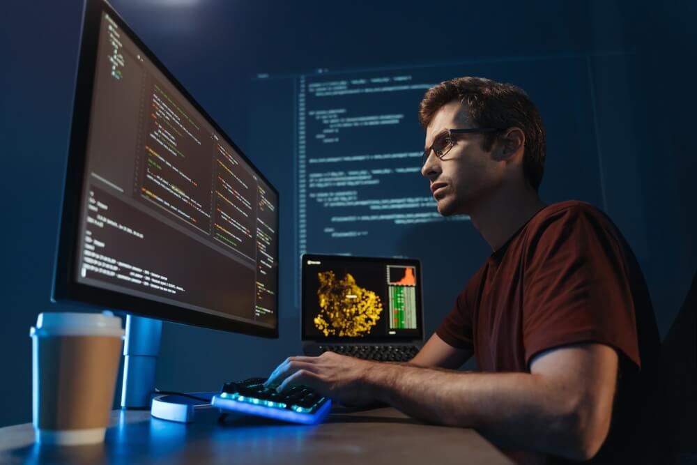 AI Web Development_Side view portrait of modern IT developer using computer at home office at night, working on software, coding new application, sitting in front of big pc monitor. Data science specialist at work