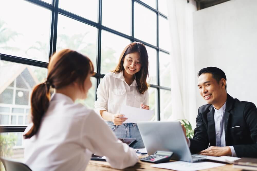 fractional marketing_Asian business woman present and explain work to female colleague, using laptop computer in office, Teamwork, coworker cooperation, financial marketing team, or corporate business concept.