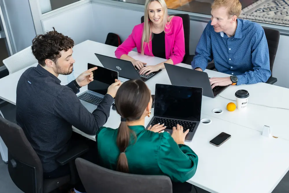 Four colleagues seated around a desk with laptops, collaborating and smiling during a meeting.