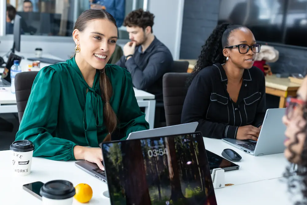 Group of colleagues working at laptops in an office, with two women in the foreground engaged in conversation.
