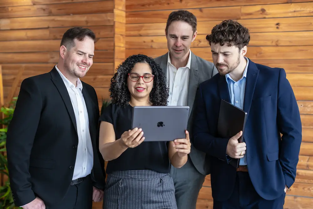 Group of four professionals in business attire smiling at the camera, two holding tablets, standing in front of a wooden wall.